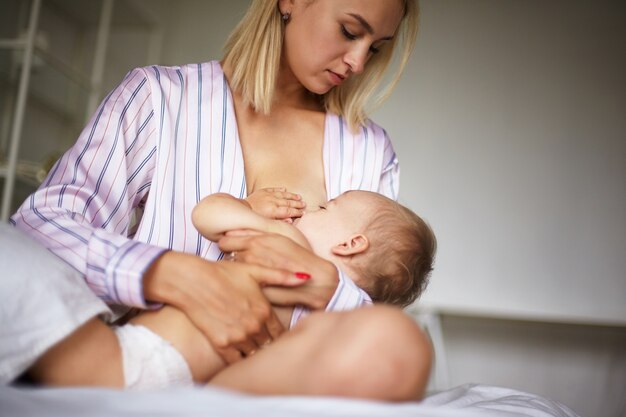 Czuła piękna młoda kobieta ubrana w stylową jedwabną piżamę siedzi na łóżku, obejmując swoje dziecko, karmiąca piersią. Śpiący maluch ssący mleko matki w sypialni. Opieka nad dziećmi, miłość i troska