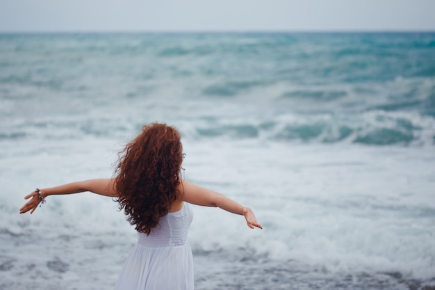 dama stoi patrząc na ręce sewith szeroko otwarte w długiej białej sukni na plaży w ciągu dnia.