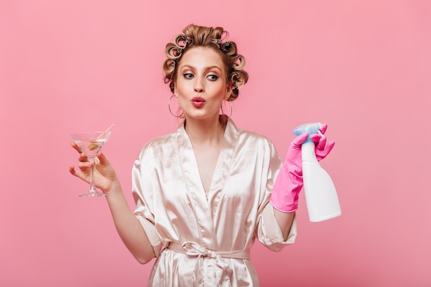 Dama w jedwabnej szacie gwiżdże, trzymając detergent i kieliszek martini na różowej ścianie