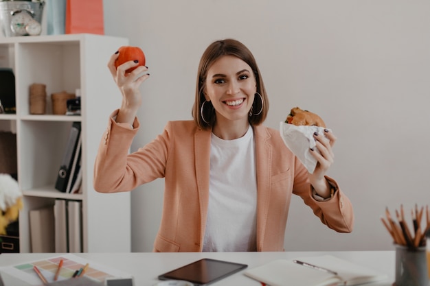 Dama w stylowym, lekkim stroju prezentuje swój lunch składający się z jabłka i burgera w miejscu pracy.