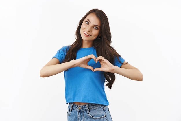 Delikatna, kobieca brunetka w basian niebieskiej koszulce uśmiechnięta i pokazująca gest serca