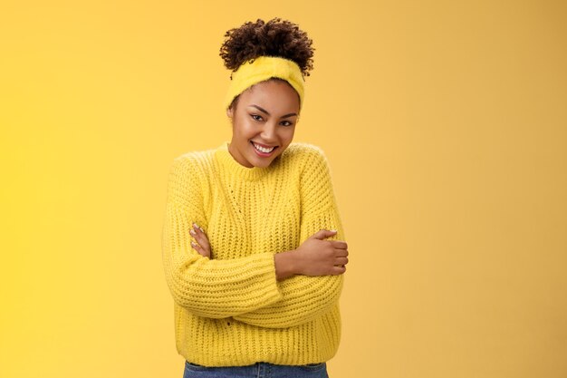 Delikatna młoda afro-amerykańska wesoła dziewczyna kręcone fryzury z pałąkiem na głowę sweter przytulanie się obejmująca radośnie uśmiechnięta kamera czuć się miękko wygodnie, stojąc na żółtym tle ciepły.