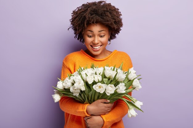 Delikatna, pozytywna Afroamerykanka obejmuje czule białe kwiaty, uśmiecha się delikatnie i patrzy na tulipany, nosi pomarańczowy sweter