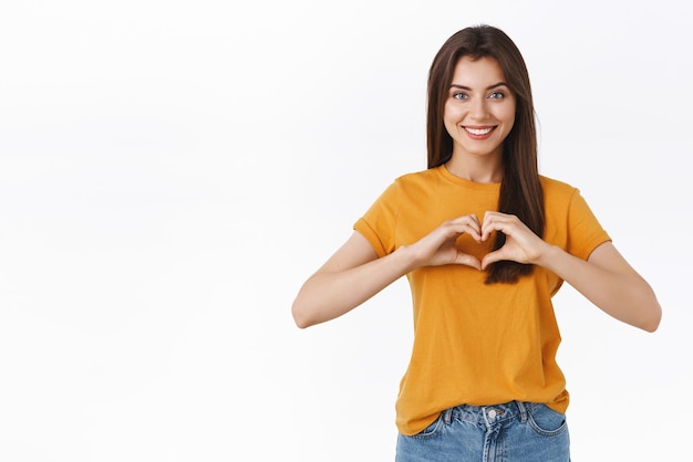 Delikatna Romantyczna I Szczęśliwa Brunetka W żółtej Koszulce Pokazująca Znak Serca Lub Miłości I Uśmiechnięta Miłosna Relacja Wyznać Z Uczuciem Lub Podziwem Stojąc Na Białym Tle Wesoła