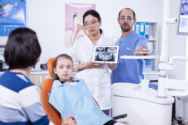 Dentysta dziecięcy omawiający profilaktykę próchnicy z rodzicem małej dziewczynki pokazującym zdjęcie rentgenowskie na tablecie. lekarz stomatolog wyjaśniający diagnozę zębów matce dziecka w przychodni na zdjęciu rentgenowskim.