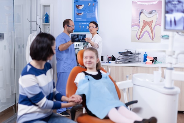 Dentysta i asystent w gabinecie dentystycznym trzyma małą dziewczynkę radiogram szczęki omawiając diagnozę. dziecko z matką podczas sprawdzania zębów ze stomatolog siedzi na krześle.