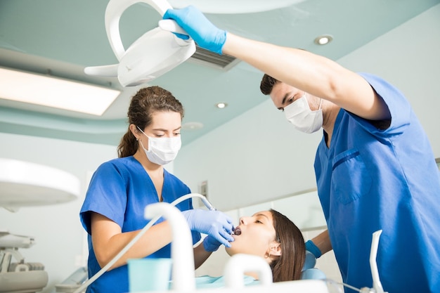 Dentysta leczący pacjenta za pomocą narzędzia dentystycznego, podczas gdy kolega dostosowuje światło w klinice