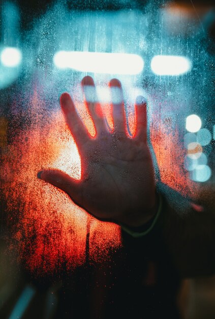Dłoń osoby dotykająca szkła pokrytego kroplami deszczu ze światłami bokeh