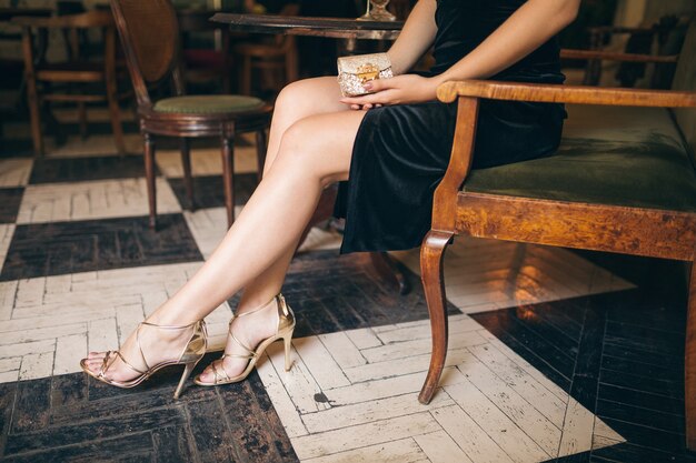 Długie chude nogi w sandałach na wysokim obcasie, modne detale eleganckiej pięknej kobiety siedzącej w kawiarni vintage w czarnej aksamitnej sukience, bogata stylowa dama, eleganckie obuwie