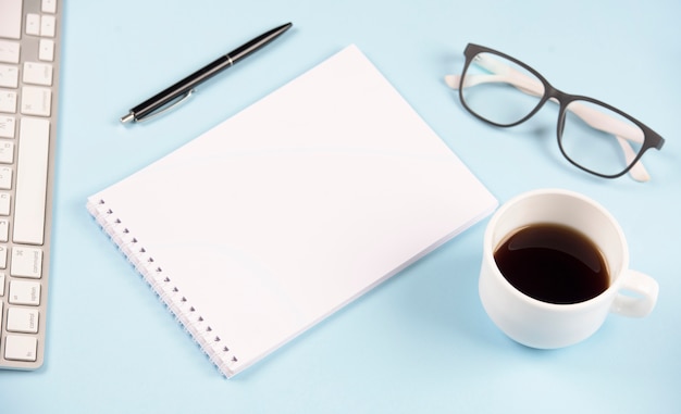 Długopis; okulary; filiżanka kawy; klawiatura i pusty ślimakowaty notepad na błękitnym tle