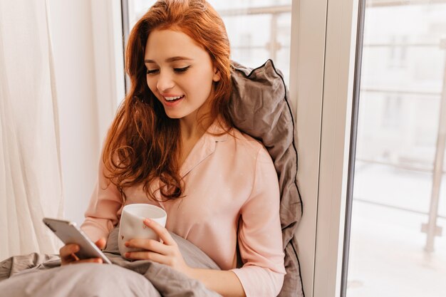 Długowłosy rudy kobieta czytająca wiadomość telefoniczną rano. Ładny kaukaski dziewczyna siedzi na łóżku z filiżanką kawy i smartfonem.