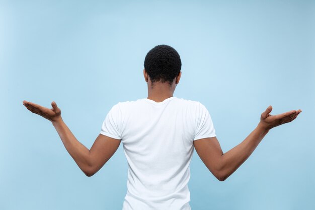 Do połowy długości z bliska portret młodego Afro-amerykańskiego modelu męskiego w białej koszuli na niebieskiej ścianie. Ludzkie emocje, wyraz twarzy, koncepcja reklamy. Wątpliwości, pytanie, niepewność.