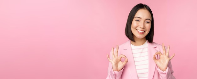 Bezpłatne zdjęcie dobra, znakomita kobieta w firmowym garniturze pokazująca ok gest aprobaty, polecająca coś, dając pozytywną opinię i uśmiechnięta, zadowolona, pozująca na różowym tle