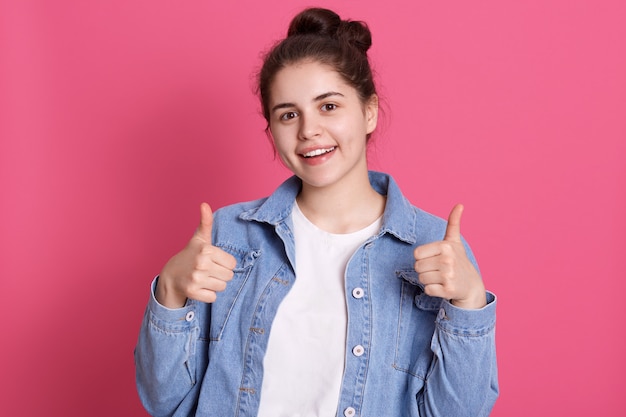 Dobrze wyglądająca szczęśliwa młoda dziewczyna nosi stylowy strój, pokazuje kciuki do góry, wyrażając pozytywne emocje