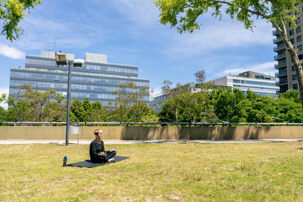 Dojrzała kobieta siedzi w pozycji lotosu ćwiczy jogę w publicznym parku w słoneczny dzień