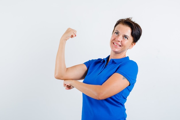 Dojrzała kobieta w niebieskiej koszulce pokazuje mięśnie ramion i wygląda pewnie, widok z przodu.