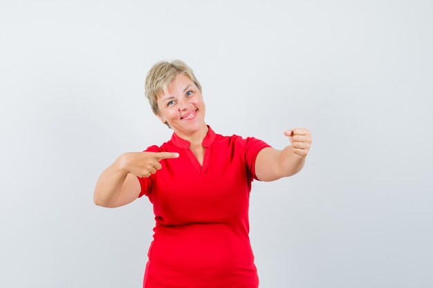 Dojrzała kobieta wskazująca na bok, udająca, że trzyma coś w czerwonej koszulce i wyglądająca wesoło