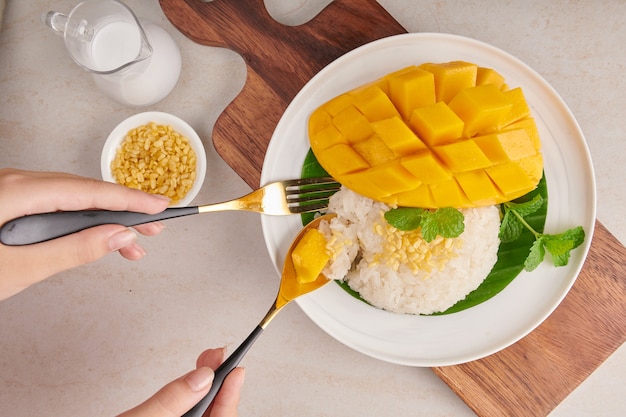 Dojrzałe mango i lepki ryż z mlekiem kokosowym w talerzu na kamiennej powierzchni, tajski słodki deser w sezonie letnim. kobieta ręce widelcem i łyżką, jedzenie mango i lepkiego ryżu. widok z góry.