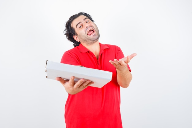 Dojrzały mężczyzna trzyma pudełko po pizzy podczas rozciągania ręki w pytającym geście w czerwonej koszulce i patrząc szczęśliwy, widok z przodu.