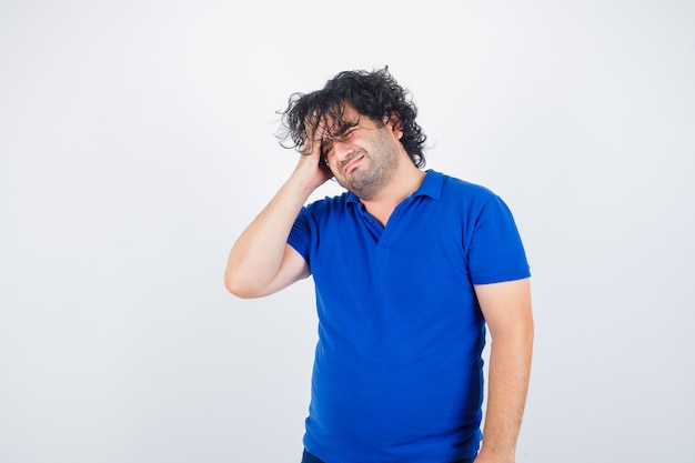Dojrzały mężczyzna w niebieskiej koszulce cierpi na silny ból głowy i wygląda na zirytowanego, widok z przodu.