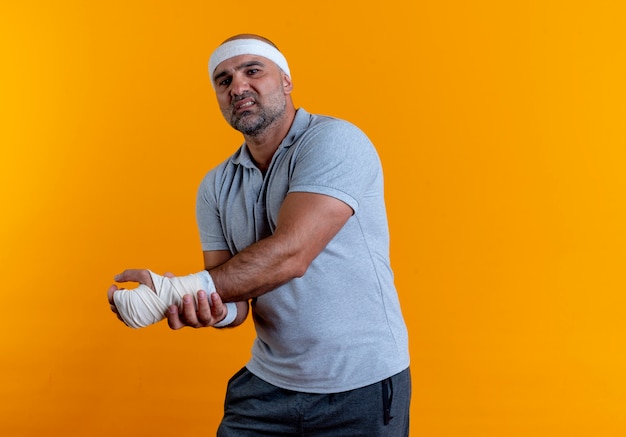 Dojrzały sportowy mężczyzna w opasce dotykając jego ręki źle wyglądający mając ból stojąc nad pomarańczową ścianą