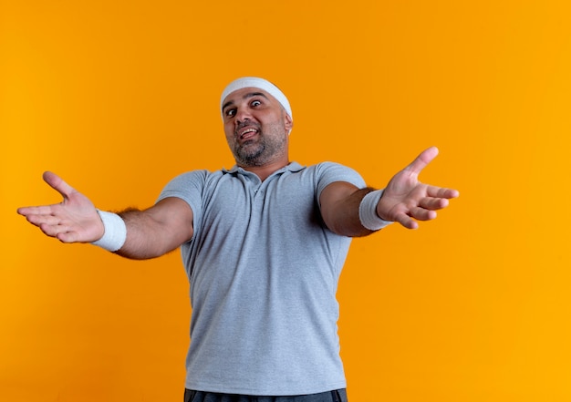 Dojrzały Sportowy Mężczyzna W Opasce Patrząc Do Przodu Robi Powitalny Gest Rękami Stojącymi Na Pomarańczowej ścianie
