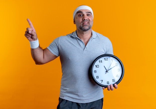Bezpłatne zdjęcie dojrzały sportowy mężczyzna w opasce trzyma zegar ścienny, wskazując palcem w bok, patrząc zdezorientowany stojąc na pomarańczowej ścianie