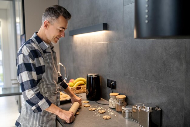 Domowe gotowanie uśmiechnięty mężczyzna stojący bokiem do kamery rozwalający surowe ciastka w domu w kuchni