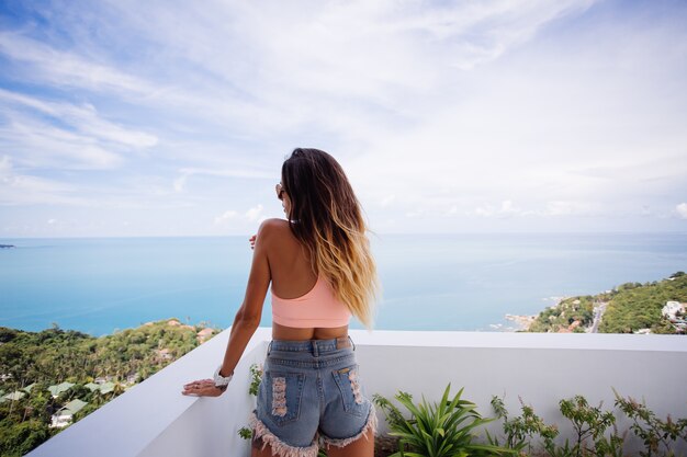 Dopasuj opaloną kaukaską kobietę w sportowym topie i spodenkach dżinsowych na balkonie luksusowej willi z tropikalnym widokiem na ocean, ciesząc się wakacjami, pozując do kamery