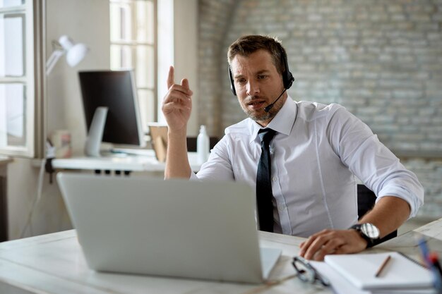 Dorosły biznesmen w średnim wieku noszący słuchawki podczas rozmowy wideo przez laptop w biurze