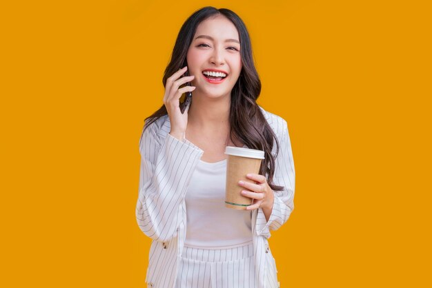 Dorywczo inteligentna azjatycka kobieta biznesu ręka za pomocą smartfona trzymaj filiżankę kawy rozmowa biznesowa uśmiechając się spójrz na kamerę pewny siebie wesoły wyraz twarzy studio strzał żółte tło