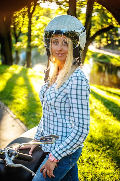 Bezpłatne zdjęcie dorywczo uśmiechnięta blond kobieta w kasku moto pozowanie na skuter na tle przyrody.