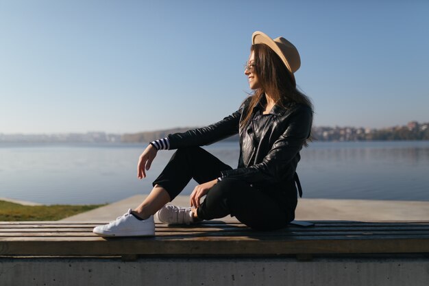 Dość młoda modelka kobieta dziewczyna pozuje siedząc na ławce w jesienny dzień na nabrzeżu jeziora
