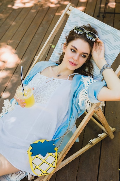 Dość młoda uśmiechnięta stylowa kobieta siedzi na leżaku w letnim stroju, ubrana w białą sukienkę, niebieską pelerynę, okulary przeciwsłoneczne, torebkę, pije świeży sok, relaks