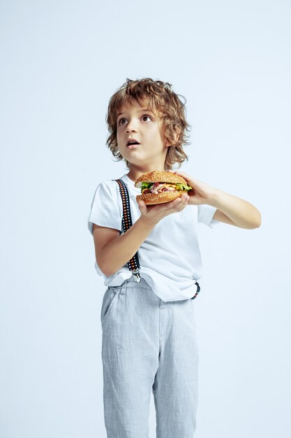 Dość młody chłopak kręcone w ubranie na białej ścianie. Jedzenie burgera. Kaukaski męski przedszkolak z jasnymi emocjami twarzy. Dzieciństwo, ekspresja, zabawa, fast food. Zdziwiony.