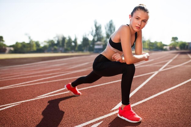 Dość sportowa dziewczyna w bezprzewodowych słuchawkach sport top i legginsy marzycielsko patrząc w kamerę i rozciągając się na bieżni stadionu miejskiego