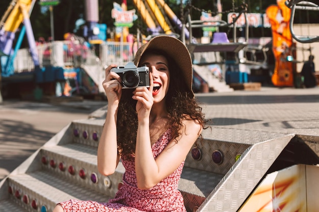 Dość uśmiechnięta kobieta z ciemnymi kręconymi włosami w kapeluszu siedzi i robi zdjęcia swoim małym aparatem, szczęśliwie spędzając czas w parku rozrywki