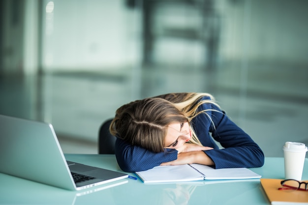 Dość wyczerpana młoda bizneswoman siedzi przy biurku do spania w swoim biurze
