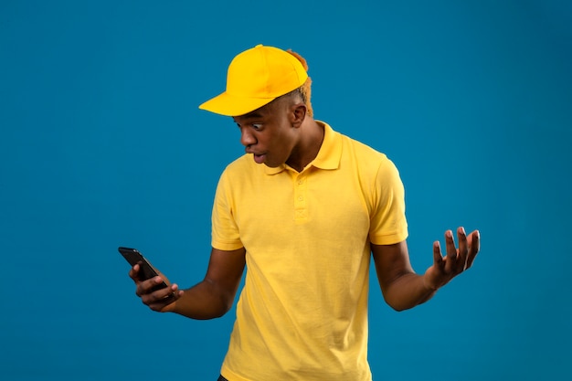 Dostarczający Afroamerykanin W żółtej Koszulce Polo I Czapce Zszokowany, Rozkładający Dłonie, Patrząc Na Ekran Oh Jego Telefon Komórkowy Stojący Na Odosobnionym Niebieskim