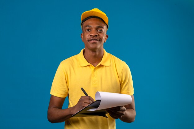 Dostawa Afroamerykanin mężczyzna w żółtej koszulce polo i czapce trzymającej schowek, piszący coś wyglądającego pewnie i dumnie stojącego na odosobnionym niebieskim