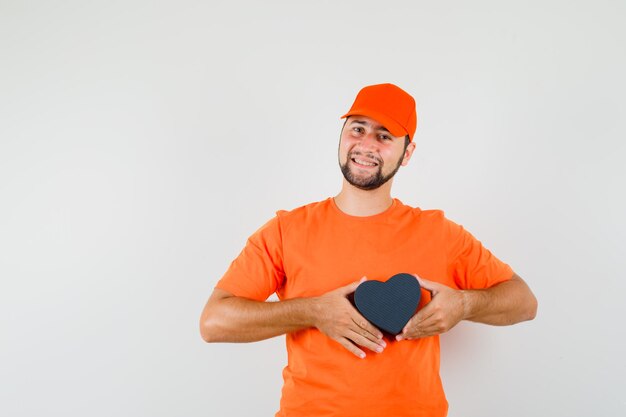 Dostawa człowiek posiadający pudełko w pomarańczowy t-shirt, czapka i patrząc wesoły. przedni widok.