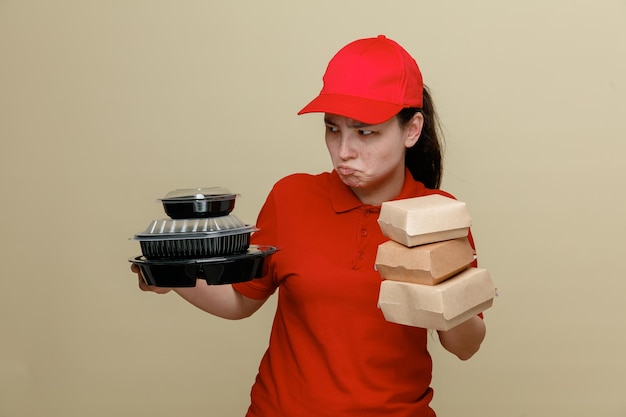 Dostawa kobieta pracownik w czerwonej czapce i pusty tshirt mundurze trzymając pojemniki na żywność patrząc na bok jest zawiedziony, marszcząc brwi, stojąc na brązowym tle
