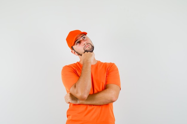 Dostawca podpierający podbródek na podniesionej pięści w pomarańczowym t-shirt, czapce i patrząc zamyślony, widok z przodu.