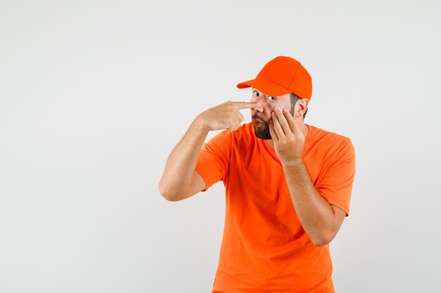 Dostawczy mężczyzna wskazujący na powiekę ściągniętą palcami w pomarańczowym t-shirt, widok z przodu czapki.