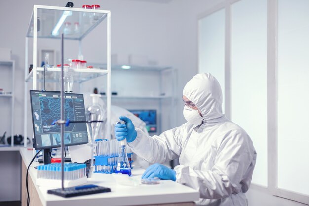 Doświadczony biolog ubrany w PPE trzymający pipetę z niebieskim roztworem. Chemik w nowoczesnym laboratorium prowadzącym badania za pomocą dozownika podczas światowej epidemii Covid-19.