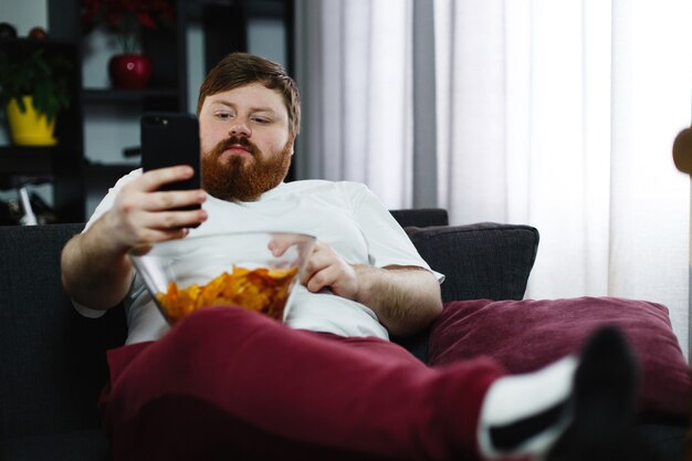 Dosyć gruby mężczyzna ono uśmiecha się sprawdzać jego smartphone podczas gdy siedzi na kanapie i je