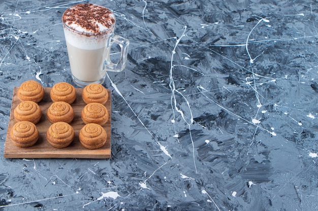 Drewniana deska słodkich okrągłych ciasteczek ze szklaną filiżanką smacznej gorącej kawy na marmurowym tle.