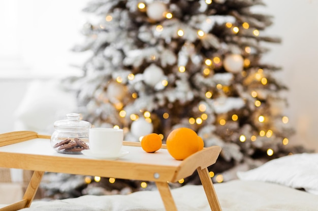 Drewniana taca z ciastkami, filiżankami i mandarynkami stoi na łóżku przed choinką ozdobioną girlandami