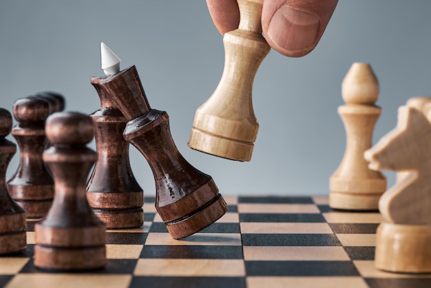 Drewniane figury szachowe na szachownicy, ręka z białą hetmanem wykonuje ruch, koncepcja strategii, planowania i podejmowania decyzji