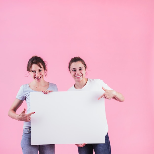 Bezpłatne zdjęcie dwa kobiety wskazuje palec nad pustą plakat pozycją przeciw różowemu tłu